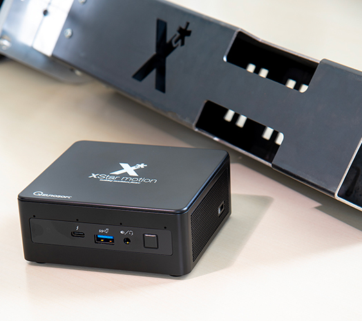 In primo piano vediamo l'XS-Box, basato su NUC intel i7 su cui è installato il server XS-Server prodotto da XStar motion. Unità plug&play che può essere interfacciata a qualsiasi macchina e ottenere così i riferimenti ottimizzati da passare alla macchina. Sul retro un tubo quadrato tagliato che riporta il logo di XStar motion.